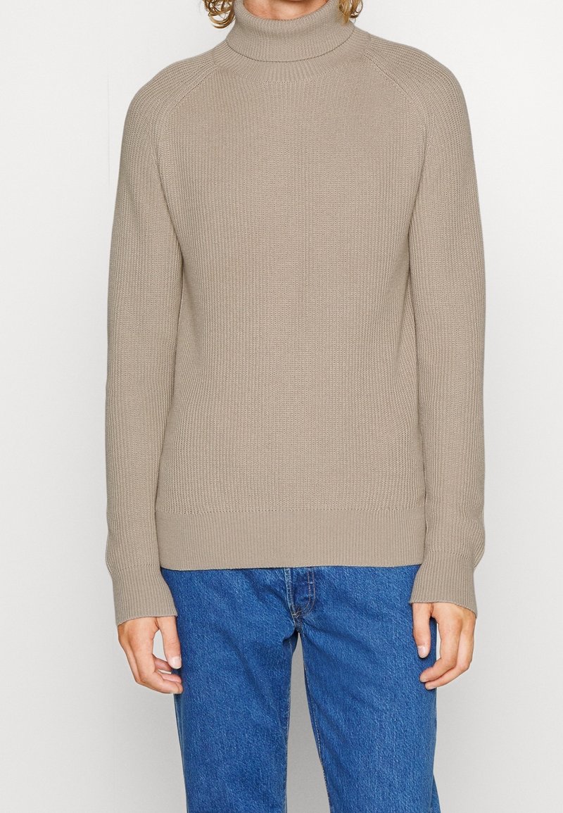 Beiger, gerippter Rollkragenpullover mit langen Ärmeln und schmalem Saum, kombiniert mit blauen Jeans. Der Pullover hat ein strukturiertes Strickmuster.
