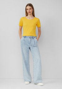 T-shirt jaune ajusté à manches courtes associé à un pantalon en denim large de couleur bleu clair. Le mannequin a les mains dans les poches, se tenant devant un fond neutre.