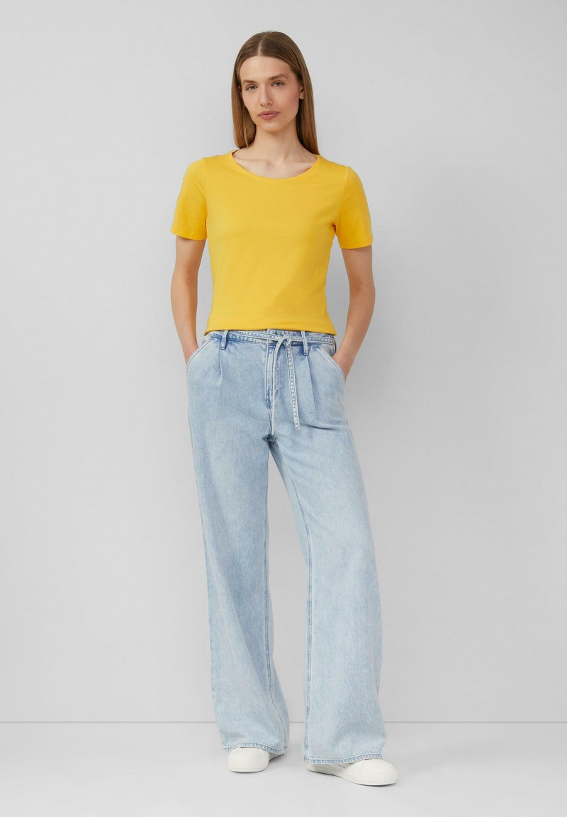 T-shirt jaune ajusté à manches courtes associé à un pantalon en denim large de couleur bleu clair. Le mannequin a les mains dans les poches, se tenant devant un fond neutre.