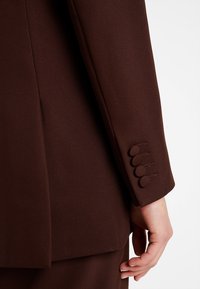 Hnědá přizpůsobená saka s jemnou texturou na rukávech, která má čtyři kulaté knoflíky na manžetě a čistý, strukturovaný design.