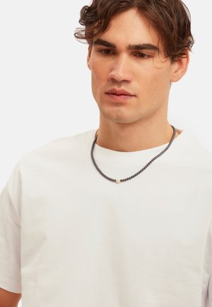 Collar de cuentas negras con una única cuenta de acento blanca, llevado sobre una camiseta blanca sencilla. El collar es corto y ajustado al cuello.