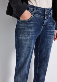 Jean en denim bleu foncé avec un délavage clair et une coupe droite. Comprend des poches avant et une taille avec une fermeture à bouton. Tissu texturé visible.