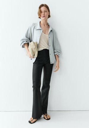 Femme portant une veste gris clair par-dessus un top beige avec un pantalon noir évasé et des sandales noires, tenant une pochette beige, debout contre un mur blanc.