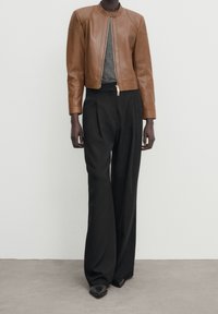 Person med en brun läderjacka över en grå topp, svarta vida byxor och svarta spetsiga skor mot en helt vit vägg.