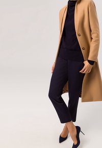 Mujer con abrigo color camello sobre un jersey negro de cuello alto, pantalones negros hasta el tobillo y tacones altos negros, de pie contra un fondo liso.