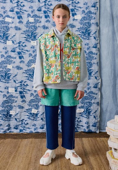 Młoda osoba stoi na tle niebieskiego, wzorzystego tła, ubrana w kwiecistą kamizelkę na szarej bluzie z kapturem, turkusowe szorty na granatowych spodniach oraz białe buty.