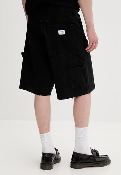 Personne posant avec un short cargo noir arrivant aux genoux, doté de grandes poches, des chaussettes blanches et des mocassins noirs sur un fond uni.