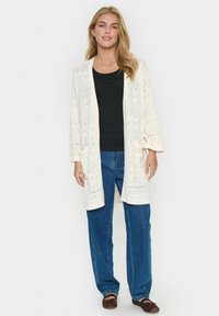 Krämsfärgad öppenstickad cardigan med 3/4-långa ärmar, med intrikata mönster; kombinerad med en svart topp och blå vida jeans.