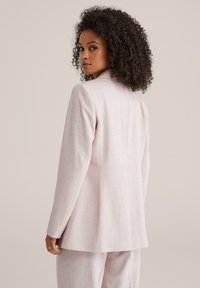 Šviesiai rožinis pritaikytas švarkas su šiek tiek tekstūruota medžiaga. Turi įpjautus šalikus, ilgus rankoves ir laisvą kirpimą, parodytas iš nugaros.