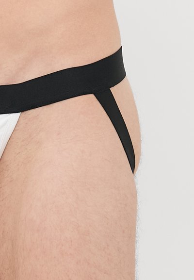 Seitenansicht einer Person, die einen schwarz-weißen Jockstrap mit einem schwarzen elastischen Bund und Bändern vor einem schlichten Hintergrund trägt.