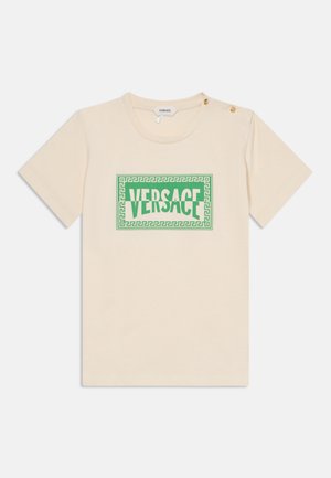 T-shirt crème à manches courtes avec logo Versace vert encadré par un motif clé grecque, et deux boutons dorés sur l'épaule gauche.