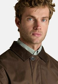 Brązowa wodoodporna kurtka z kołnierzem z materiału kordowego, zapinana na jeden guzik, noszona na zielono-białą w paski koszulę.