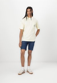 T-shirt en coton jaune avec une poche sur la poitrine et un motif graphique, associée à un short en jean et des baskets blanches avec des accents noirs.