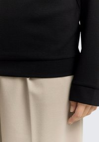 Pull noir texturé à manches longues, associé à un pantalon plissé beige, mettant en valeur un tissu lisse et une coupe décontractée. Main reposant sur la couture.