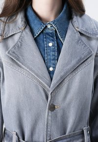 Veste en denim gris clair avec un col à revers, des coutures visibles et un accent de bouton noir. Portée par-dessus une chemise en denim bleue avec des boutons en perles.
