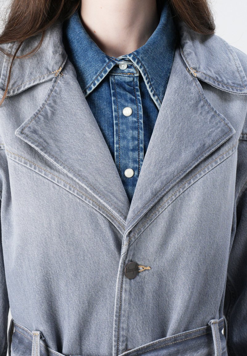 Hellgraue Jeansjacke mit Reverskragen, sichtbaren Nähten und einem schwarzen Knopfakzent. Getragen über einem blauen Jeanshemd mit Perlenknöpfen.