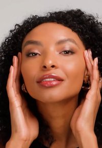 Femme aux cheveux bouclés touchant doucement son visage, portant des boucles d'oreilles en or et un maquillage naturel, sur un fond clair uni.