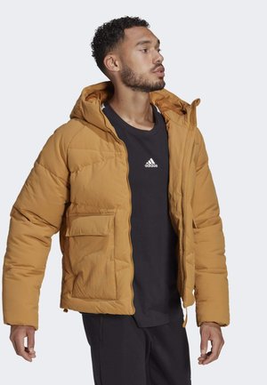 Hořčicově žlutá puffer bunda s kapucí, s předním zipovým zapínáním, bočními kapsami a mírně texturovanou tkaninou. Nositá přes černý top.