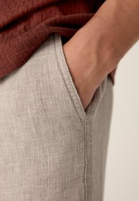 Mano descansando dentro del bolsillo de unos pantalones de tela beige con textura, usando una camiseta de textura color óxido.