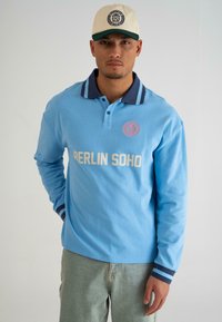 Svijetloplavi dugosleeve polo s tamnoplavim ovratnikom i prugastim manžetama. Ima natpis "BERLIN SOHO" ispisan bijelom bojom na prsima. Ležeran dizajn.