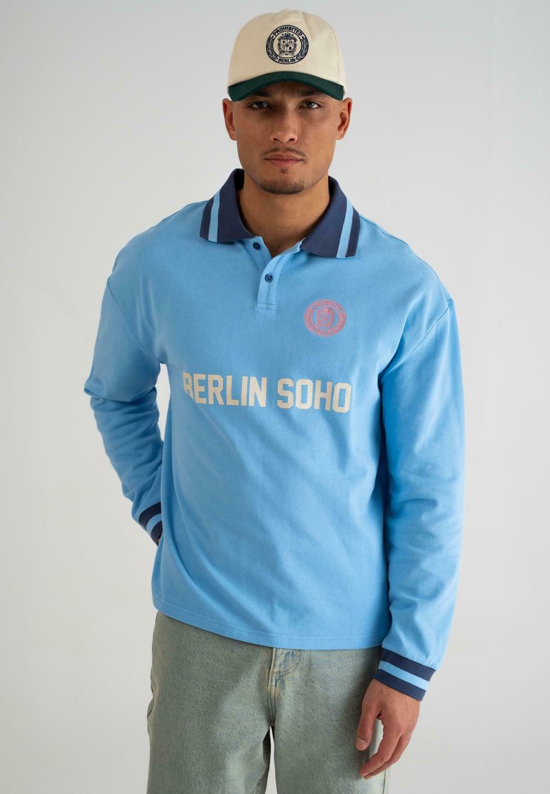 Svijetloplavi dugosleeve polo s tamnoplavim ovratnikom i prugastim manžetama. Ima natpis "BERLIN SOHO" ispisan bijelom bojom na prsima. Ležeran dizajn.