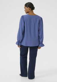 Blue, long-sleeve blouse with puffed shoulders and gathered cuffs, paired with dark blue flared jeans, viewed from the back.