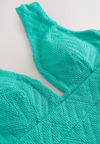 Gros plan d’un tissu turquoise texturé avec des motifs tricotés en zigzag et ondulés formant des sections structurées.
