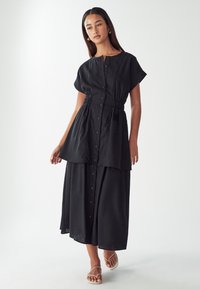 Abito nero con bottoni, maniche corte, vita cintrata, design a strati e gonna fluida. Il tessuto ha una texture liscia e uno stile minimalista.