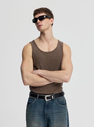 Mężczyzna w czarnych okularach przeciwsłonecznych, brązowej bezrękawniku, niebieskich dżinsach i czarnym pasku, z założonymi na siebie ramionami na tle gładkim jasnym.