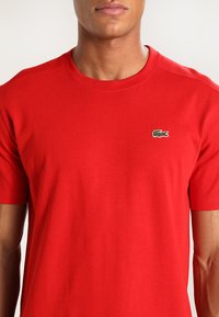 Rød bomulls-T-skjorte med korte ermer og rund hals, med en liten grønn broderte logo på venstre bryst.