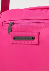 Borsa in tessuto rosa con tasca con zip e un'etichetta in metallo nero con la scritta "PENSARE FUORI DAGLI SCHEMI" impressa. Texture liscia e design durevole.