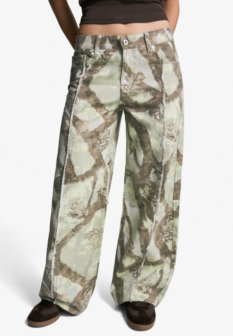 Hlače širokih nogavica v kamuflažnem vzorcu, v odtenkih zelene in rjave, izdelane iz trpežnega blaga, z vidnimi šivi in sproščenim krojem.