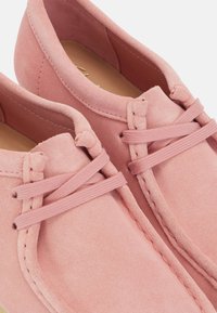 Rosa mockasiner med rund tå, dekorativa snören och synlig söm längs kanterna. Slät textur med matta ytor.