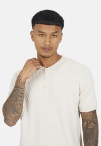 Jasno beżowa koszulka polo z teksturowanym wzorem w romby, z krótkim rękawem i kołnierzykiem z trzema guzikami. Złożone tatuaże widoczne na ramieniu.