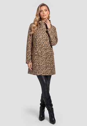 Kvinde i leopardprint langærmet frakke, sorte læderleggings og sorte slouchy hælede støvler står med den ene hånd nær ansigtet.