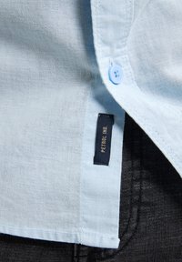 Cămașă din in albastru deschis, cu închidere cu nasturi, având un etichetă din material etichetată "PETROL INC." lângă materialul denim închis.