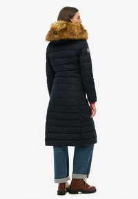 Gewatteerde marineblauwe jas tot de knie met een gefesselde capuchon van bont, horizontaal geborduurd ontwerp en een bontaccent. Gecombineerd met opgerolde spijkerjeans en bruine laarzen.