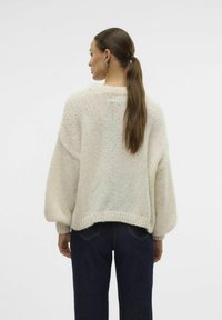 Pull en tricot crème, épais, avec une coupe ample, des manches chauves-souris et un ourlet côtelé. Porté sur un jean en denim foncé, mettant en valeur un style décontracté.