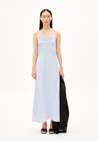 Robe maxi en satin bleu clair avec des fines bretelles, un décolleté carré, une silhouette fluide, portée avec une veste noire et des sandales à glissière noires élégantes.