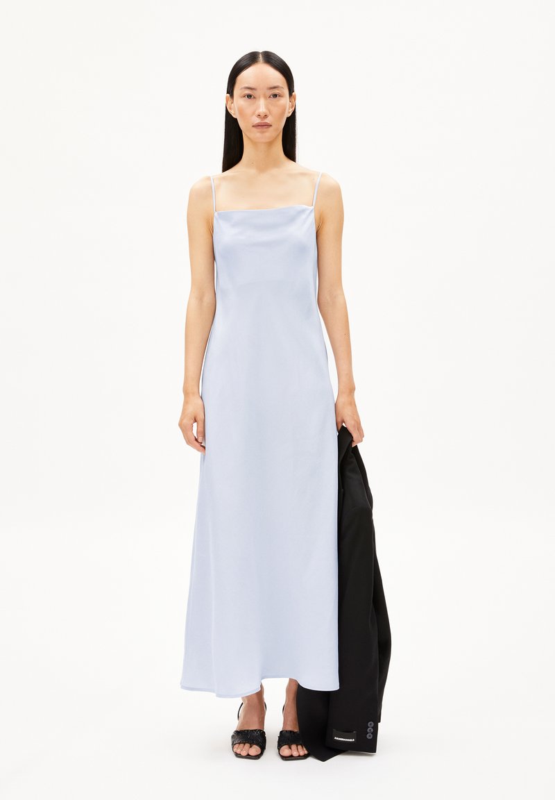 Robe maxi en satin bleu clair avec des fines bretelles, un décolleté carré, une silhouette fluide, portée avec une veste noire et des sandales à glissière noires élégantes.