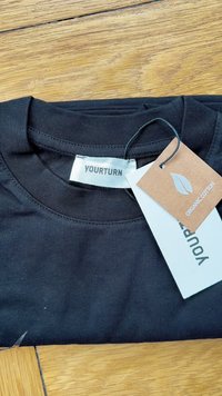 T-shirt preto em algodão orgânico com decote redondo. A etiqueta apresenta a marca "YOURTURN" e uma etiqueta castanha indicando "ALGODÃO ORGÂNICO" com um design de folha.