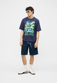 Νέος άνδρας με νεον πράσινα γυαλιά ηλίου, μπλε μπλούζα Puma με γραφικό και λουλουδάτο σχέδιο, σκούρο τζιν σορτς, λευκές κάλτσες και λευκά αθλητικά παπούτσια.