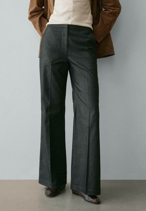 Persona che indossa pantaloni ampi sartoriali grigio scuro, scarpe in pelle marrone, maglia beige e giacca in camoscio marrone con le mani nelle tasche.