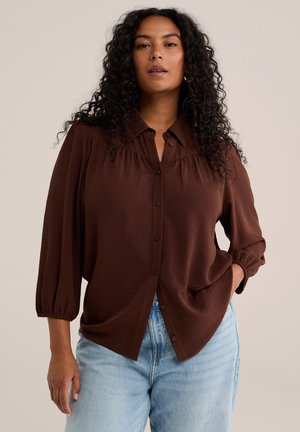 Woman with curly dark hair wears a loose brown buttoned blouse with balloon sleeves and light blue jeans, posing against a neutral background.