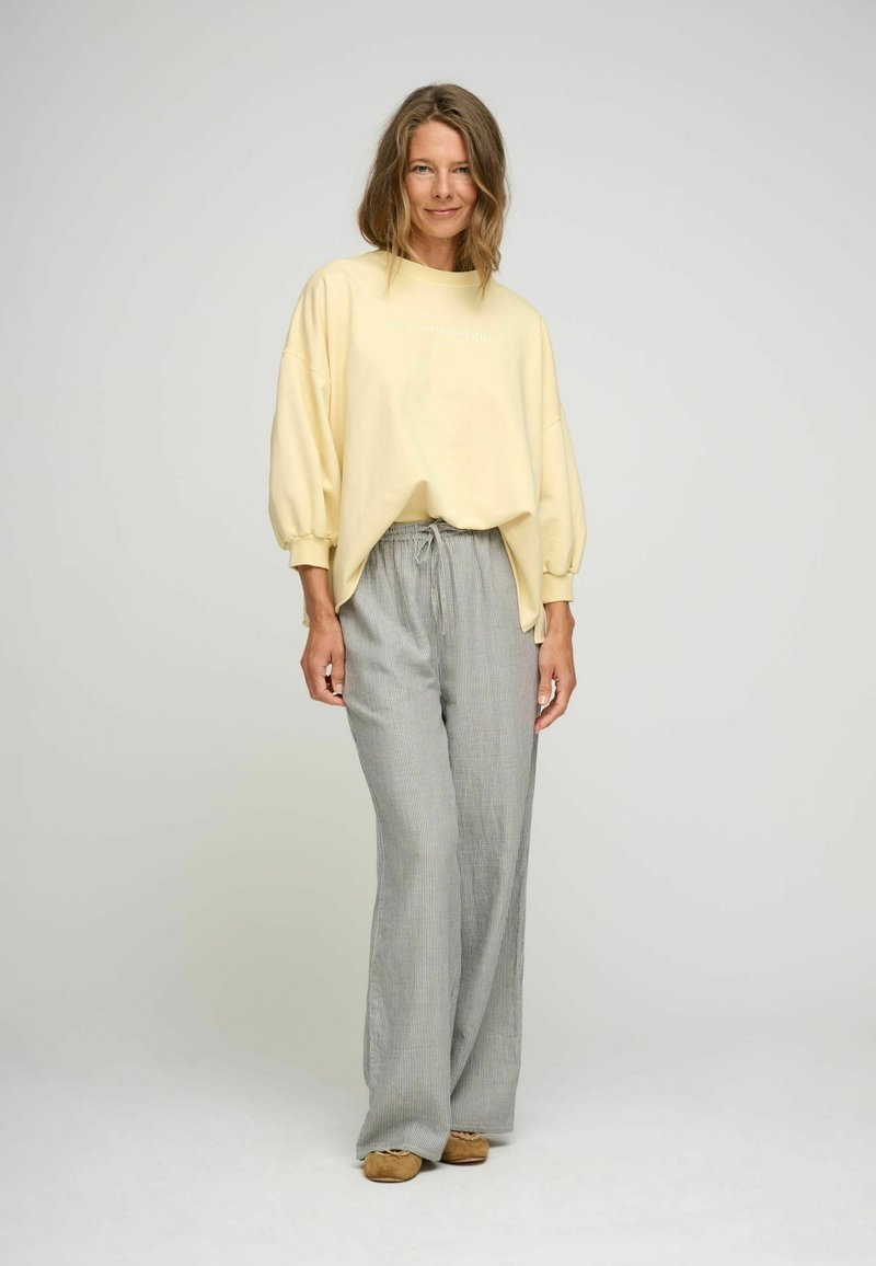 Žltá mikina s oversized rukávmi, bielym textom; šedé pruhované nohavice s širokými nohavicami; model má na sebe hnedé slip-on topánky. Jednoduché pozadie.