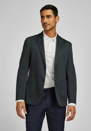 Marineblazer mit Struktur, zwei Vorderaschen, Reverskragen und zwei Knöpfen, getragen über einem weißen Poloshirt und dunklen Hosen.