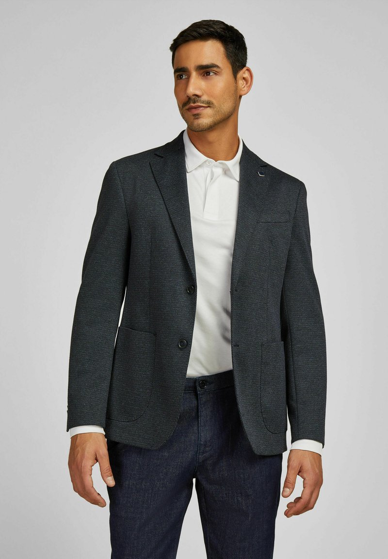 Marineblazer mit Struktur, zwei Vorderaschen, Reverskragen und zwei Knöpfen, getragen über einem weißen Poloshirt und dunklen Hosen.