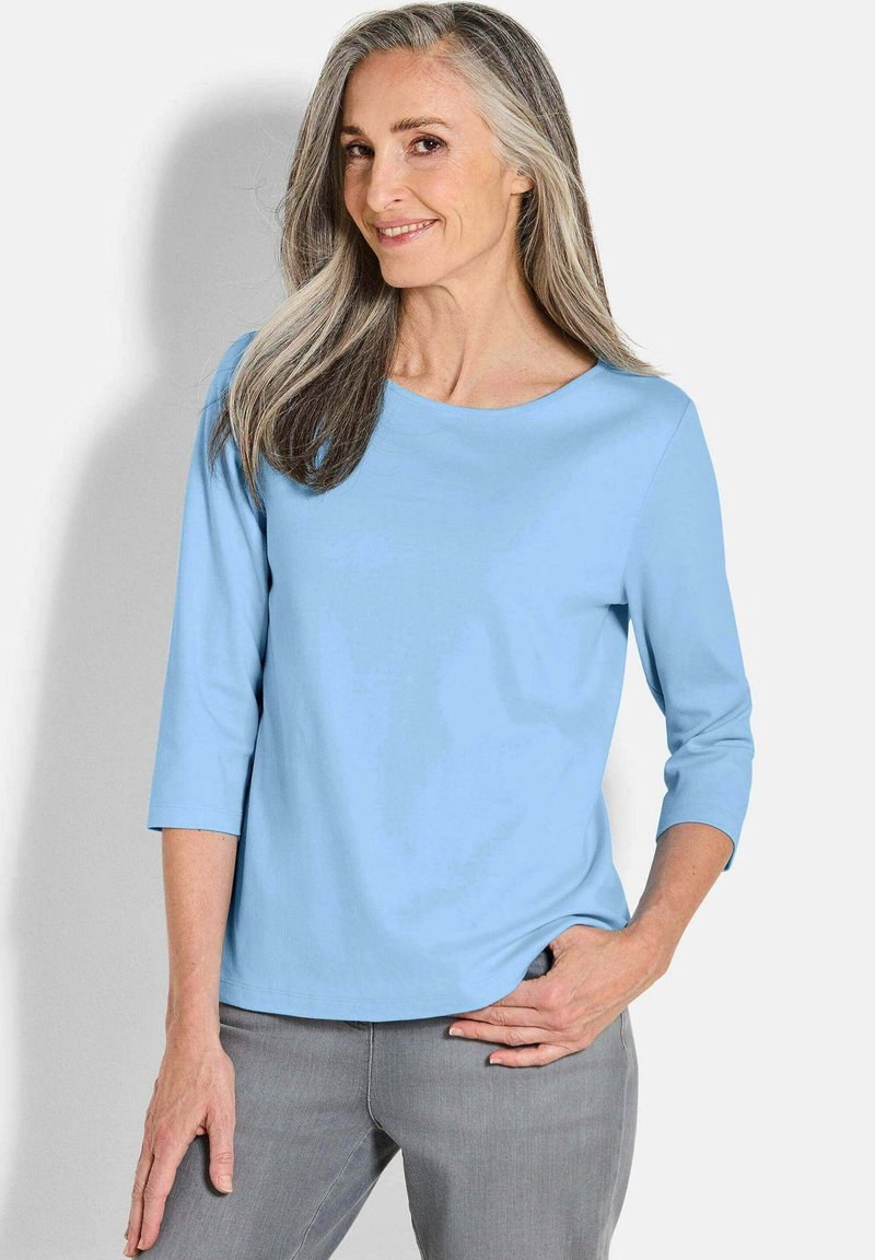 Helles blaues Oberteil mit drei Viertelärmeln aus glattem Stoff und rundem Ausschnitt. Kombiniert mit grauen Hosen, die eine lockere Passform zeigen.