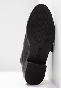 Chaussure en cuir noir avec une semelle en caoutchouc texturée, une large bride au milieu et une finition lisse avec peu de détails.