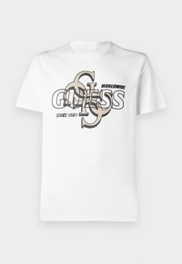 Hvid bomulds t-shirt med et grafisk design, der har overlappende tekst "GUESS" og "WORLDWIDE SINCE 1981" i sort og beige. Korte ærmer.